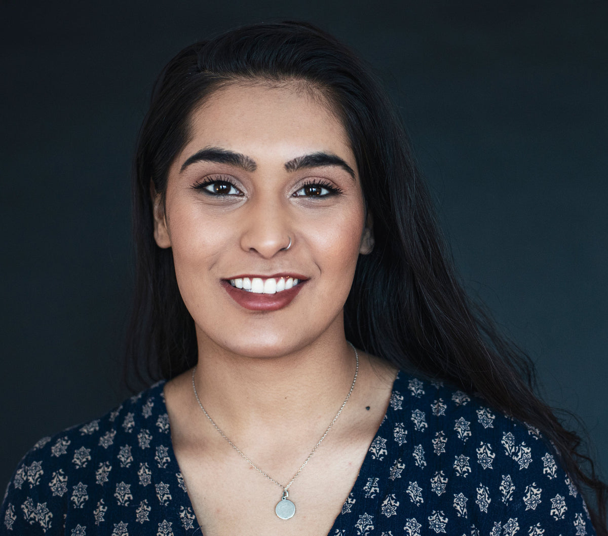 Woman with long dark hair wearing a dark blue floral top against a dark background